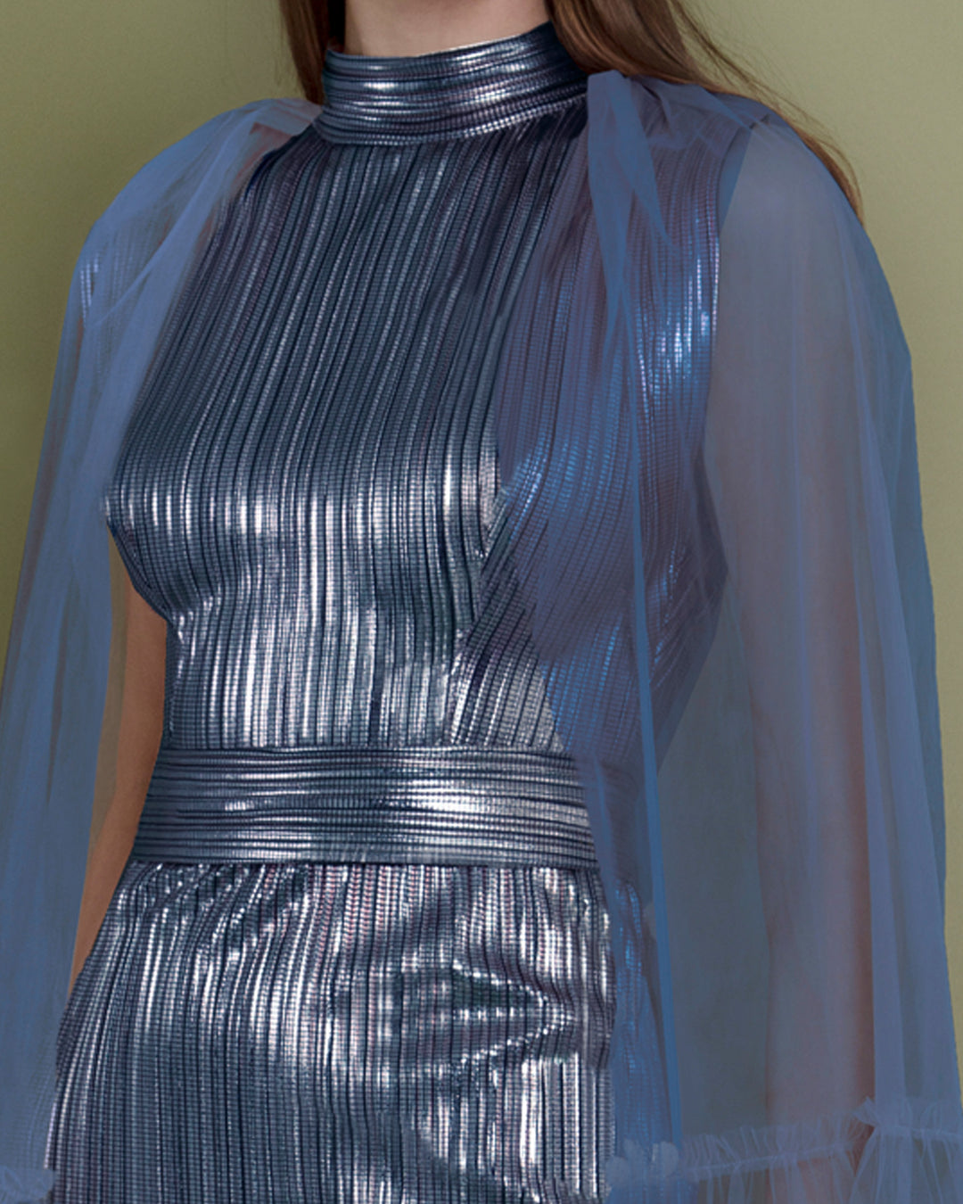 A close-up of a slim cut metallic blue evening dress in pleated fabric, paired with a tulle cape.