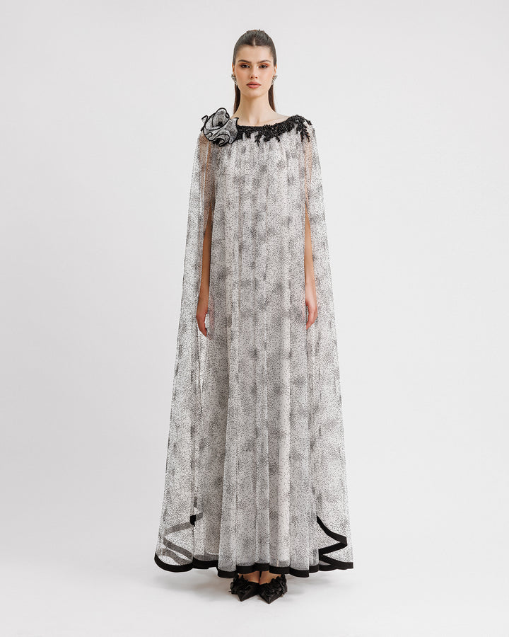 A loose-cut kaftan-style black dotted white tulle dress featuring beadings on the neckline, a draped flower on the shoulder, and a black hemline.