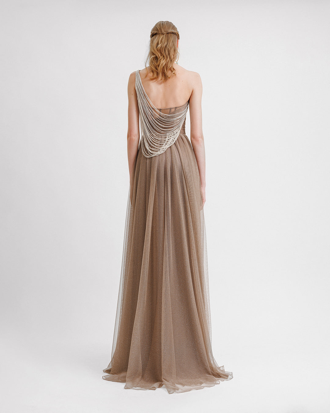 The back of a one-shoulder draped shiny tulle dress in taupe color featuring an asymmetrical tasseled back.