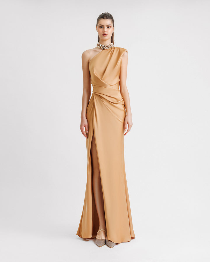 A one-shoulder draped evening dress in caramel color satin with floral embellishments on the neckline and an open slit on the side.