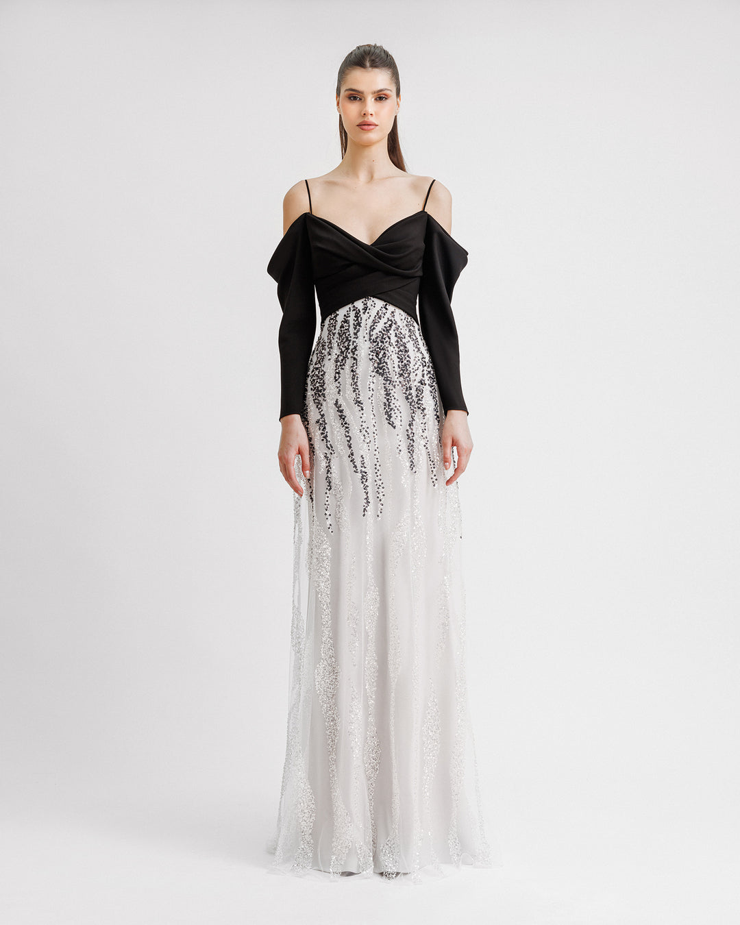 A long black and white evening dress with draped off-the-shoulder sleeves in black crepe fabric and a black and white sequins overlaying a long white skirt.