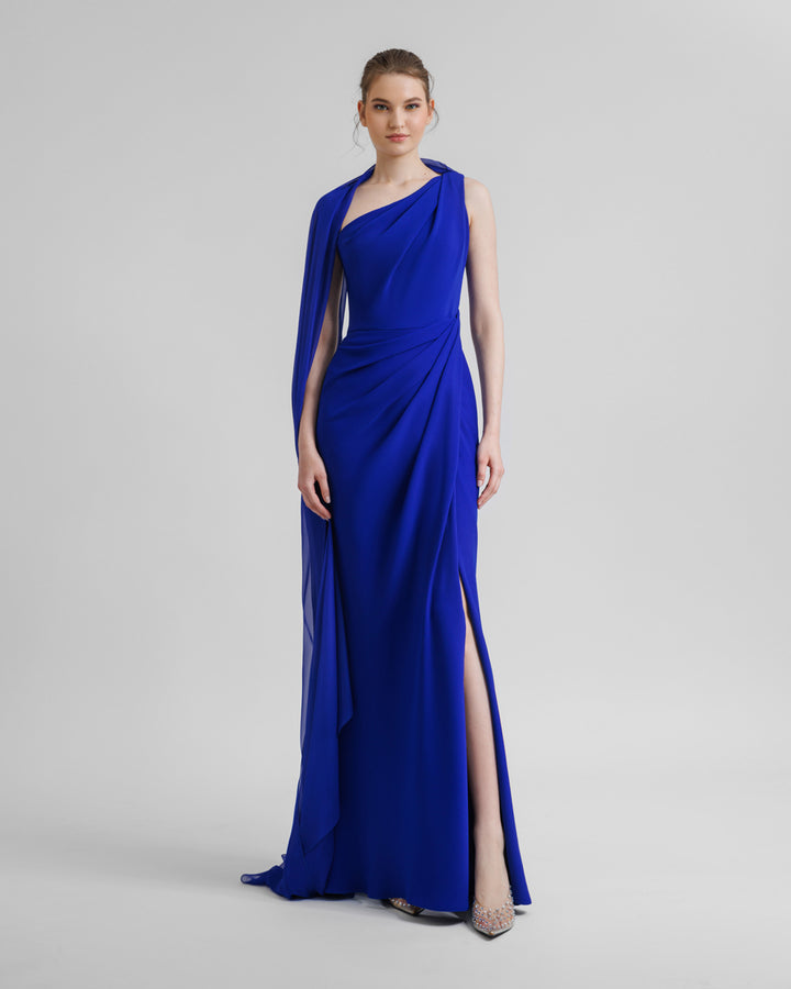 An asymmetrical floor length shawl collar royal blue evening dress with draping details on the shoulder and the waist. It features an open slit on the side.