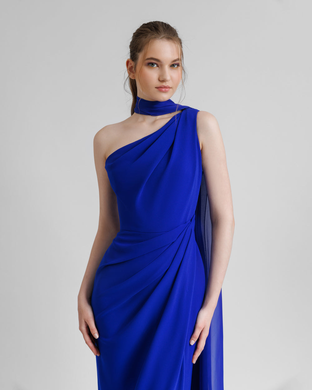 A close-up of an asymmetrical shawl collar blue evening dress with draping details on the shoulder and the waist.