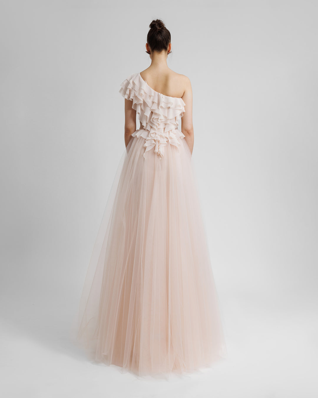 The back of a one-shoulder cut corset evening dress in blush color, with ruffled details on the bodice part, see through fabric on the waist, and a tulle skirt.