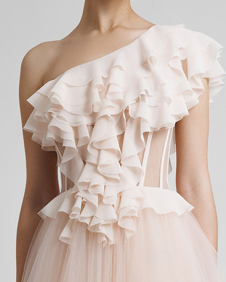 A close-up of a one-shoulder cut corset evening dress in blush color, with ruffled details on the bodice part, see through fabric on the waist, and a tulle skirt.