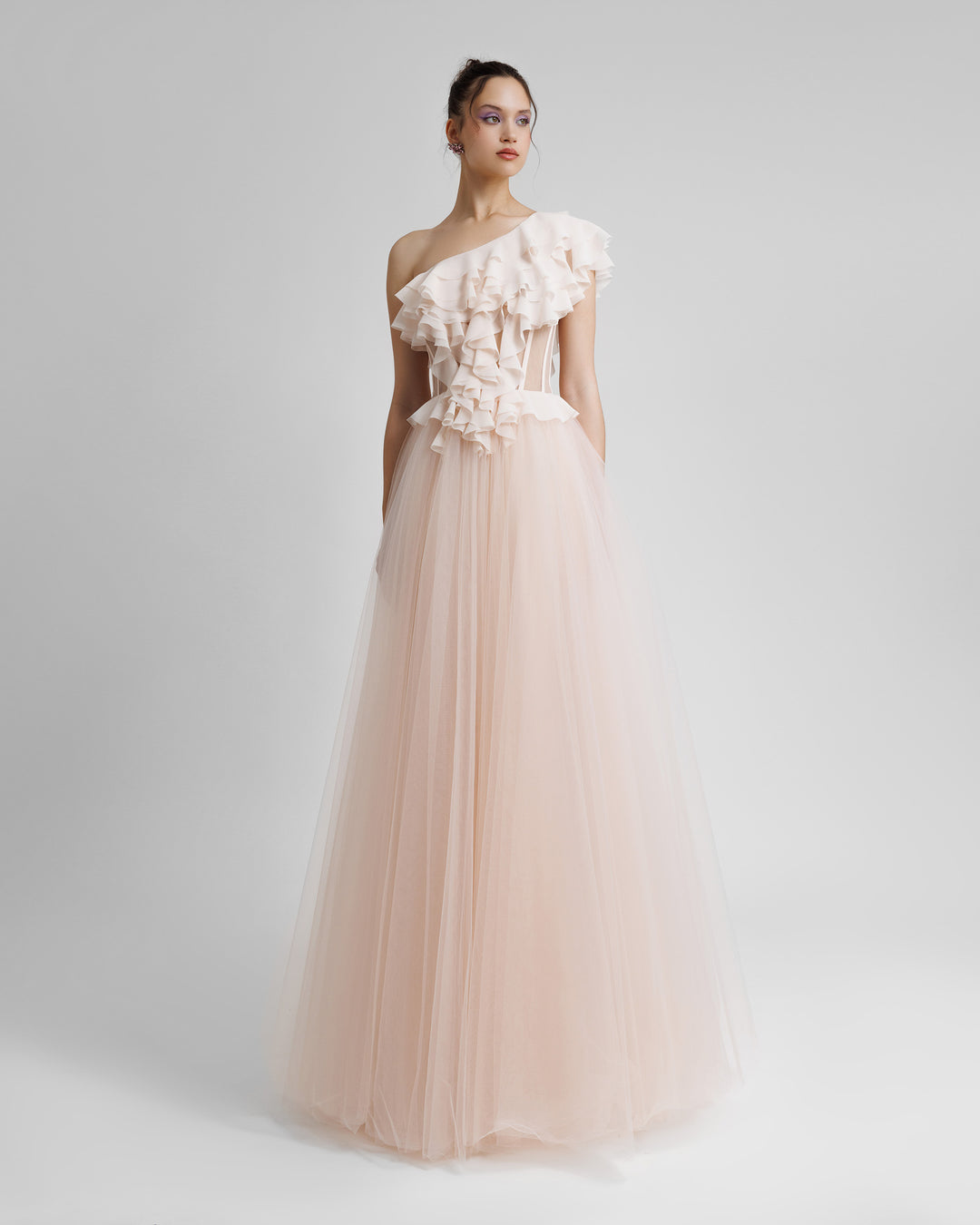 A one-shoulder cut corset evening dress in blush color, with ruffled details on the bodice part, see through fabric on the waist, and a tulle skirt.