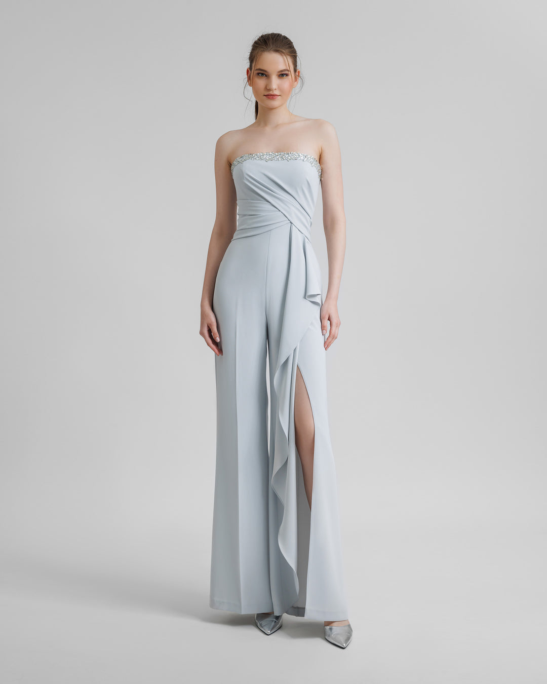 A strapless grey jumpsuit featuring a beaded neckline, draping on the waist and a ruffled slit on one leg.