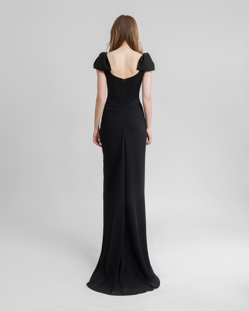 The back of a squared neckline corset long black evening dress with bow designs on the shoulders and draping details on the waist.