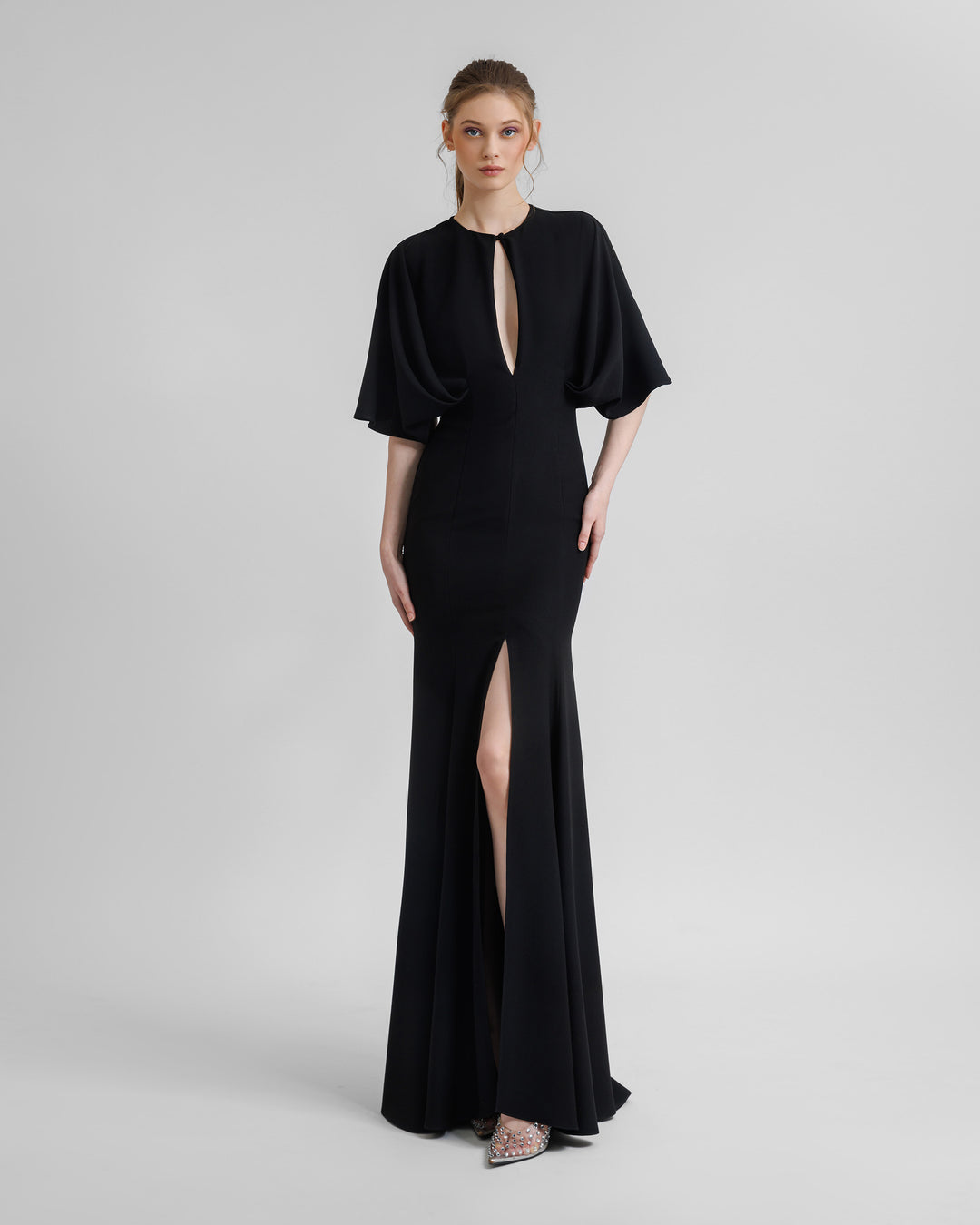 A slim-cut black evening dress with a front opening on the bust area and a middle slit, and wide cape-like sleeves.