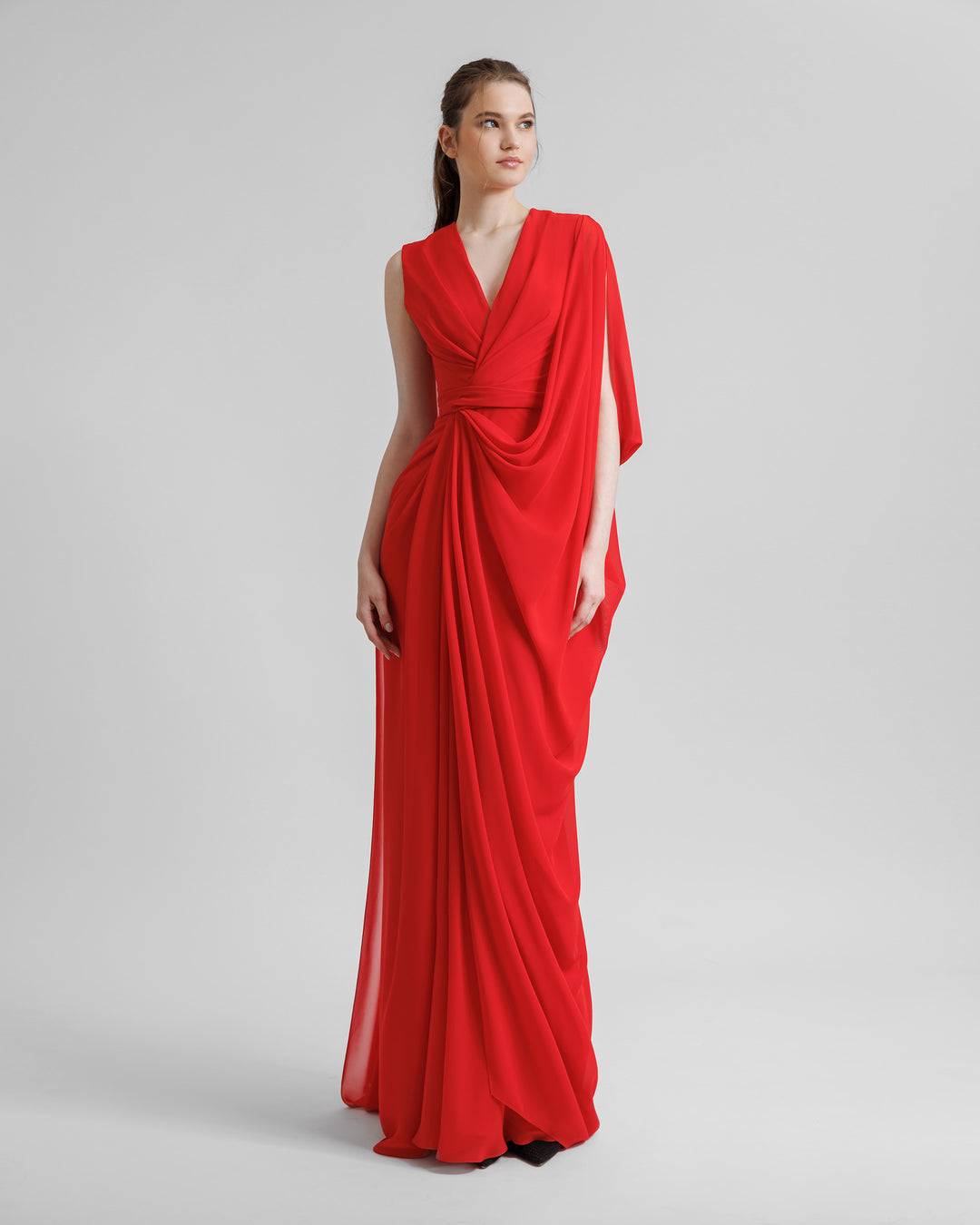 An asymmetrical V-neckline, red evening dress with draping details, and a long cape-like sleeves.