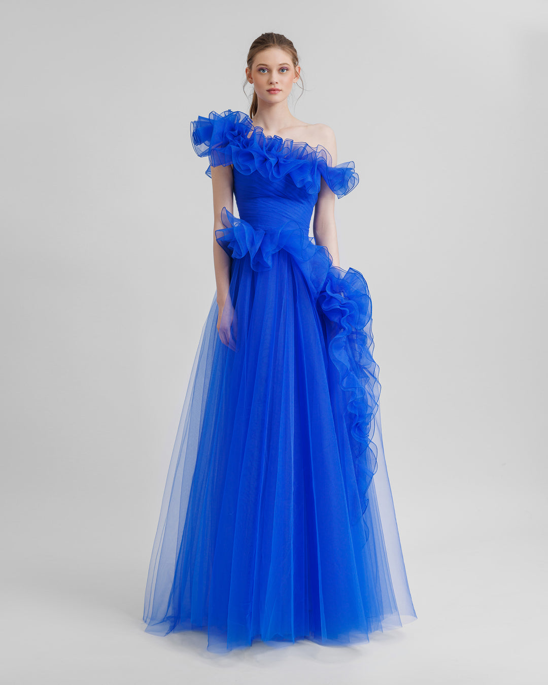 A flared royal blue evening dress featuring an asymmetrical ruffled neckline, and ruffled embellishments on the skirt.