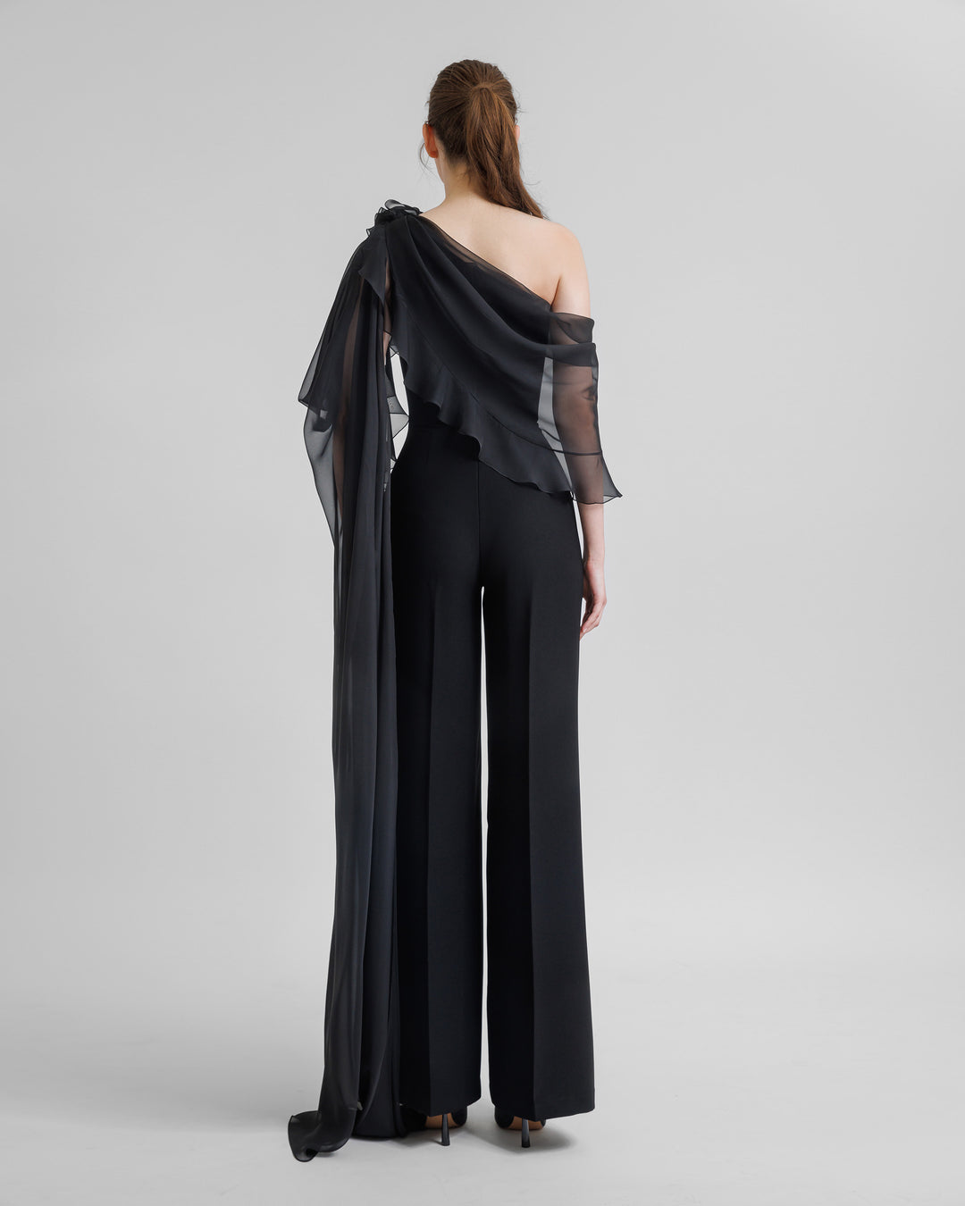 The back of a flared crepe black jumpsuit featuring asymmetrical ruffled cape-like chiffon sleeve with flowers details.