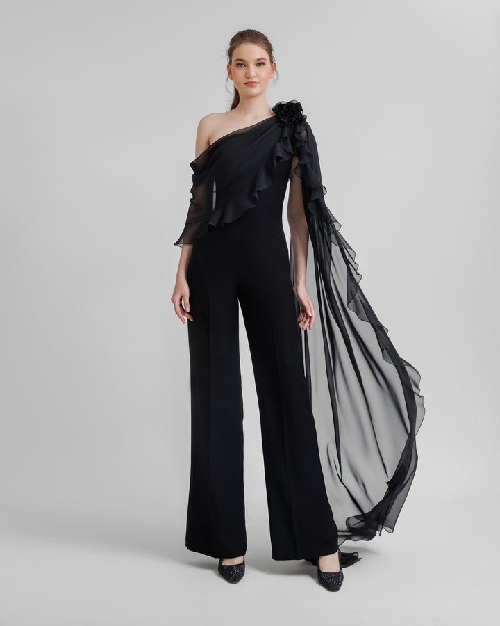 A flared crepe black jumpsuit featuring asymmetrical ruffled cape-like chiffon sleeve with flowers details.