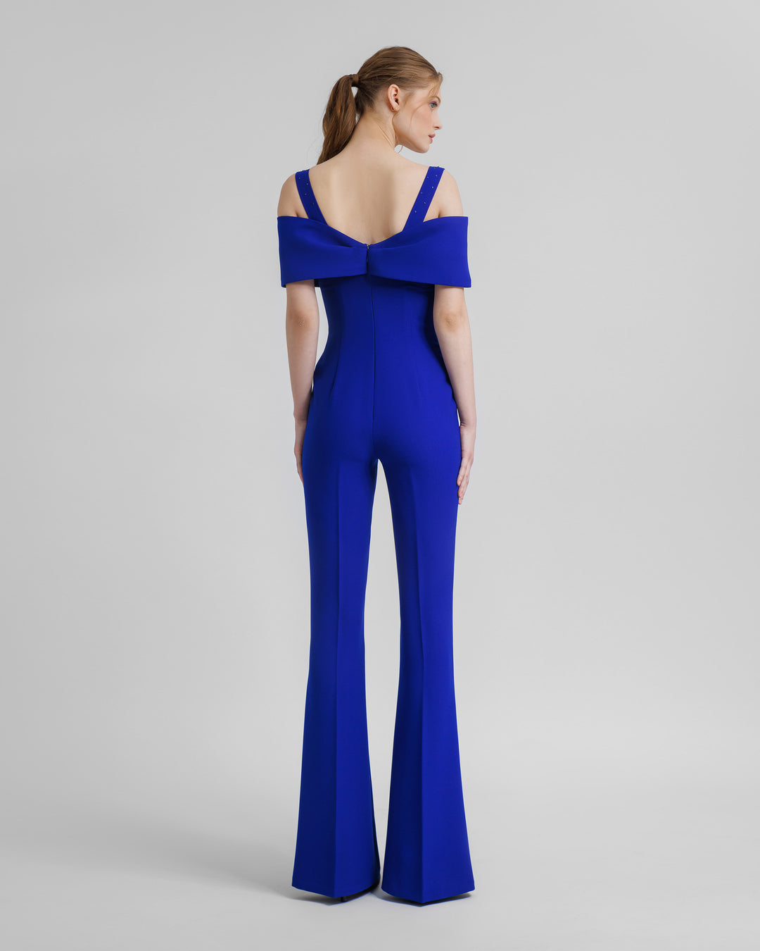 The back of a flared royal blue jumpsuit featuring an off-shoulders corset bow design and beaded straps.