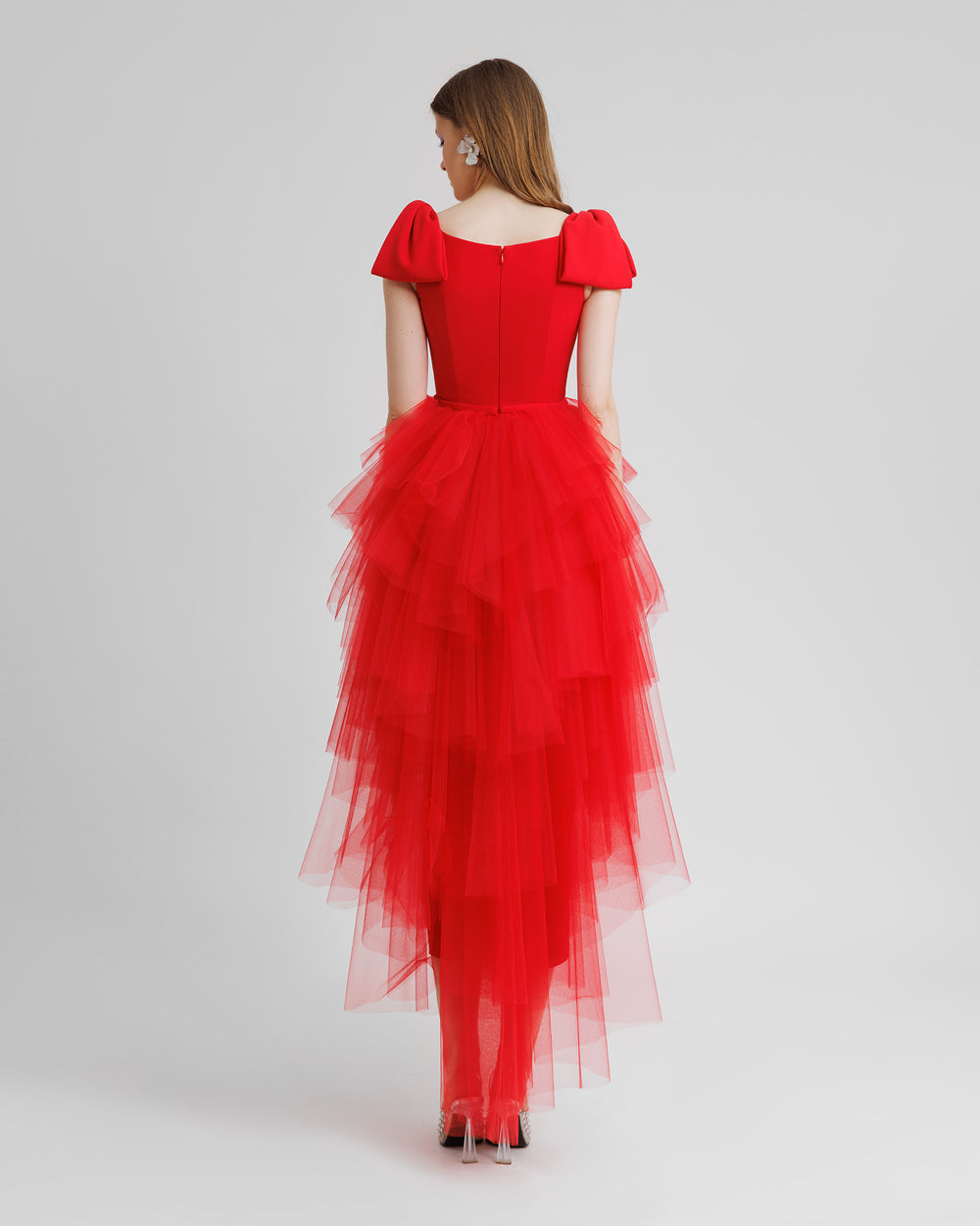 The back of a long red evening dress with bow designs on the shoulders and asymmetrical rushed tulle layered skirt.