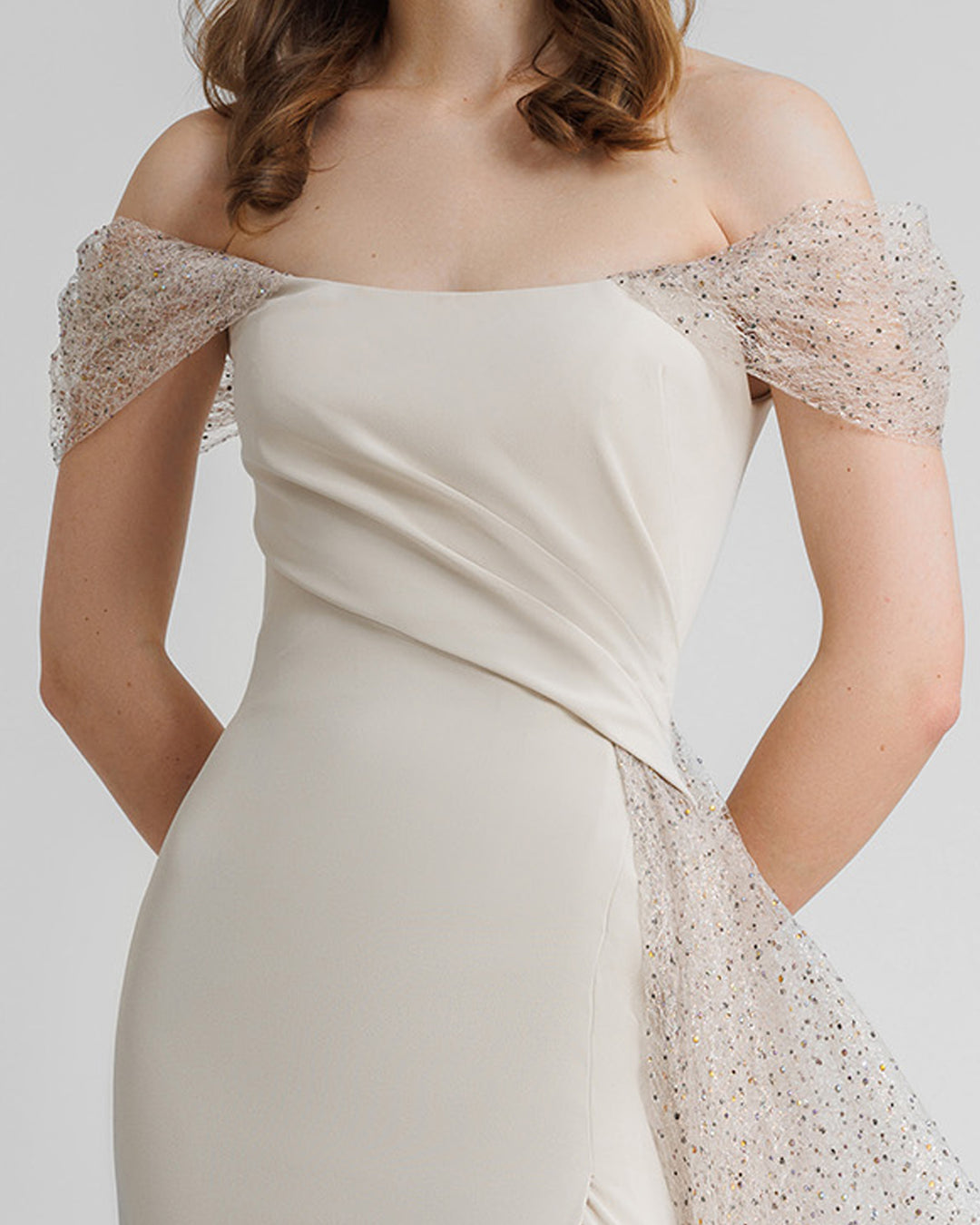 A close-up of an off-the-shoulders long champagne evening dress with draping details on the bust, and an asymmetrical side tail.