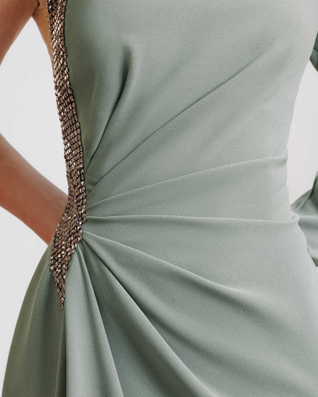 A close-up of a stone embellished evening dress in sage color with draping on the waistline.