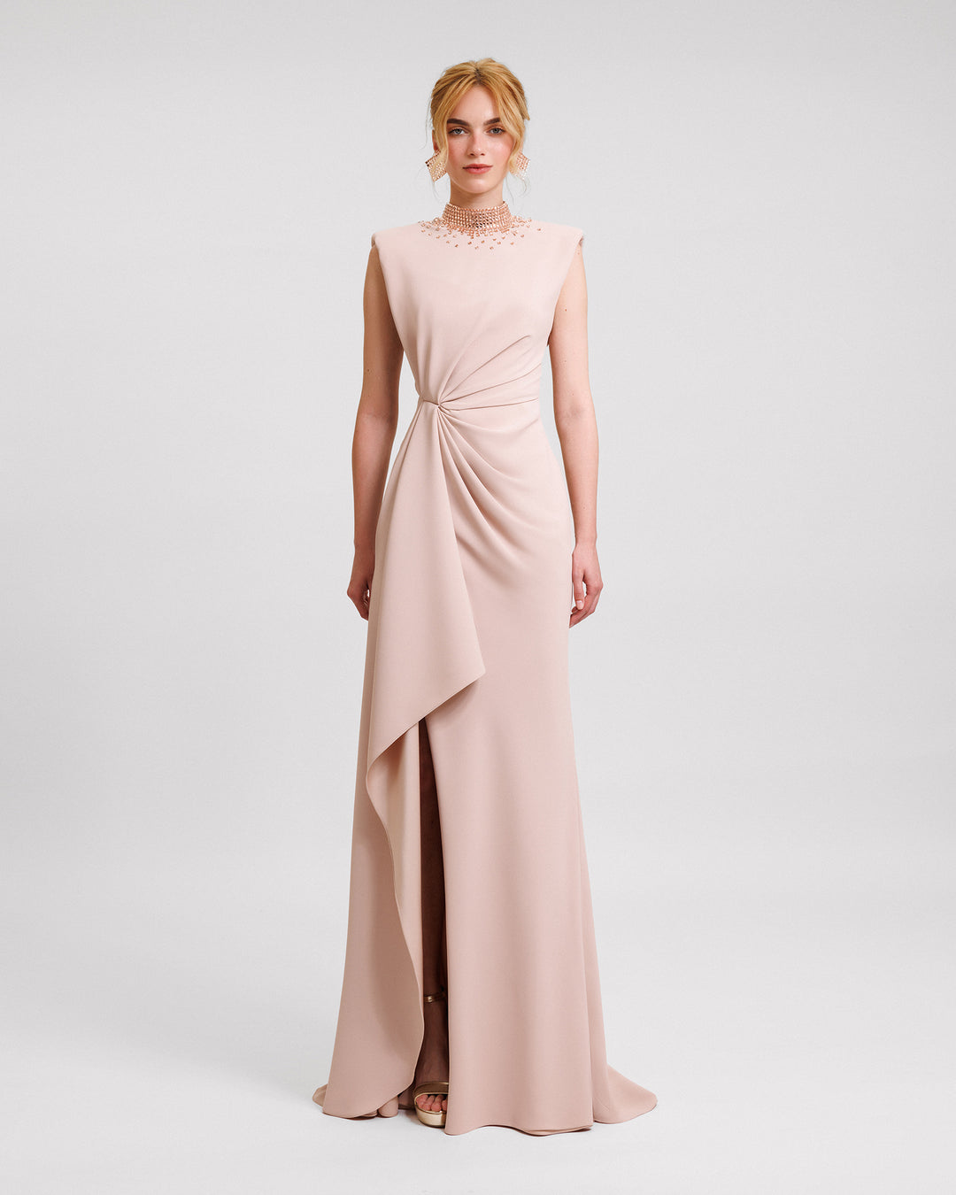 A long blush evening dress featuring a stone embellished collar, draping on the waist, a cut-out at the back, and a slit on the side.