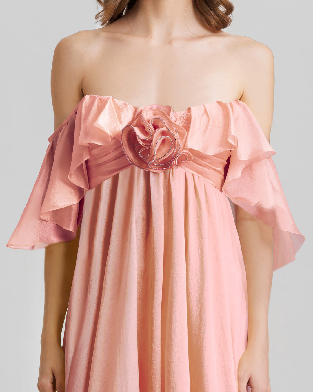 A close-up of a ruffled off-the-shoulders flowy dress in salmon color with a draped flower at the front.