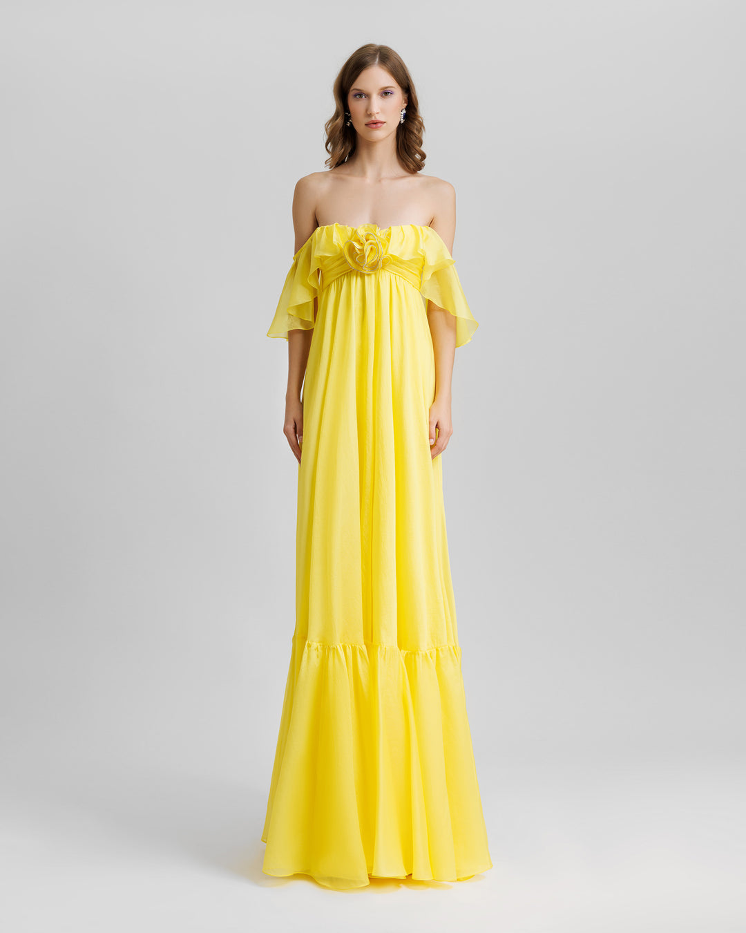 A ruffled off-the-shoulders flowy yellow evening dress with a draped flower at the front.