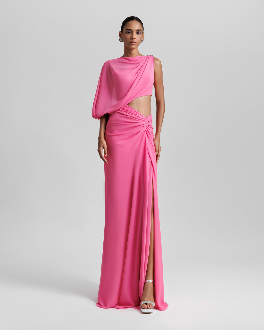 An asymmetrical pink evening dress with a draped sleeve, cut out on the waist, draped skirt, and a slit on the side.