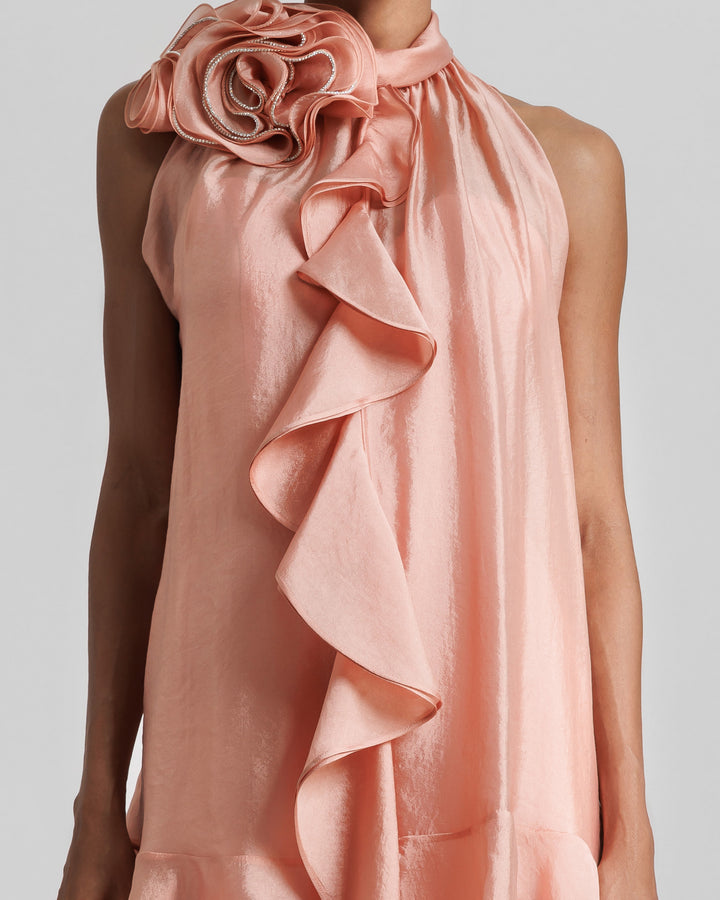 A close-up of a high-collar dress in salmon color, featuring a draped flower on the neckline and ruffles at the front.