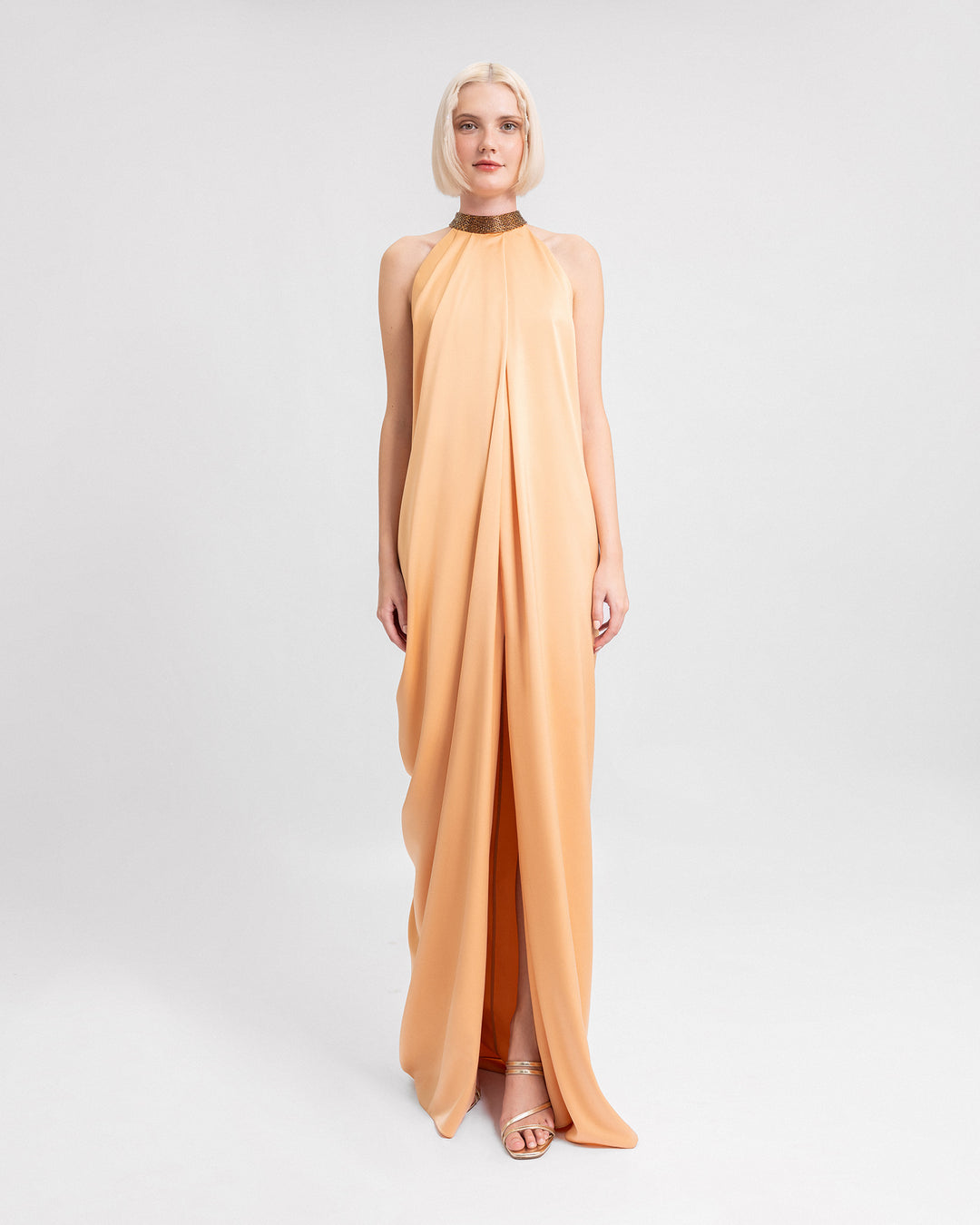 A loose-cut draped evening dress in peach satin featuring a beaded collar and a slit at the front.