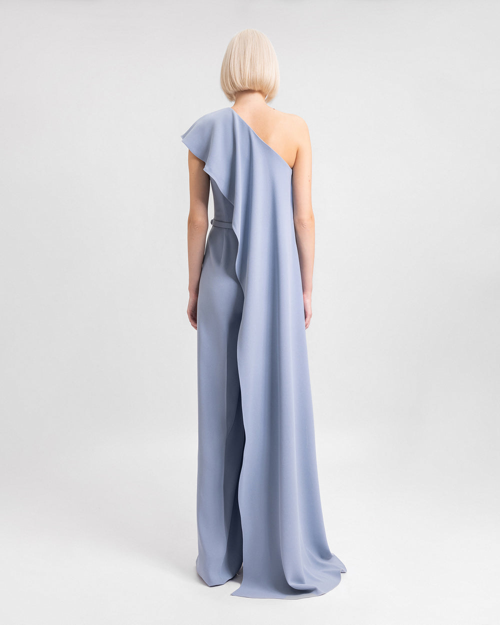 The black of a one-shoulder straight-cut jumpsuit in dusty blue with a long ruffled neckline and a detachable belt.