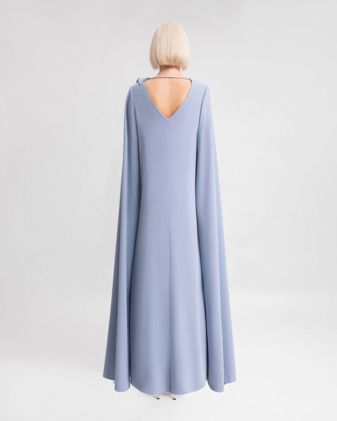 The back of a long loose-cut evening dress in dusty blue with open floor length sleeves and a triangle cut out on the back.