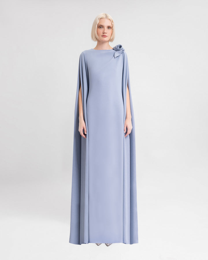 A long loose-cut evening dress in dusty blue with open floor length sleeves and a draped flower on the shoulder.