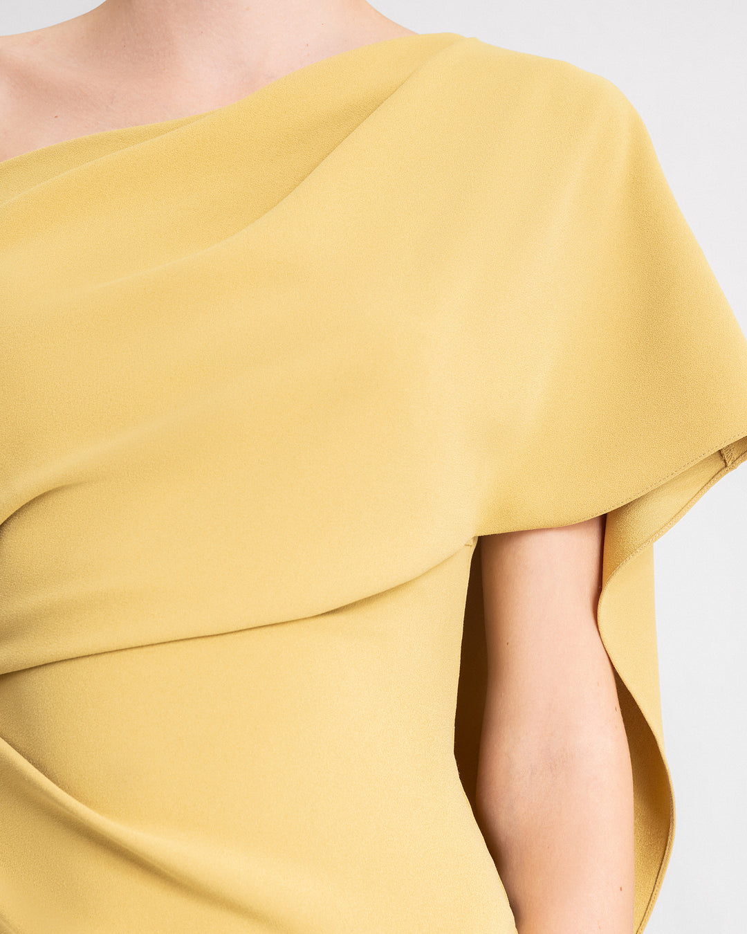 A close-up of an asymmetrical neckline evening dress in mustard color featuring draping on the waistline and a long ruffled cape-like sleeve.