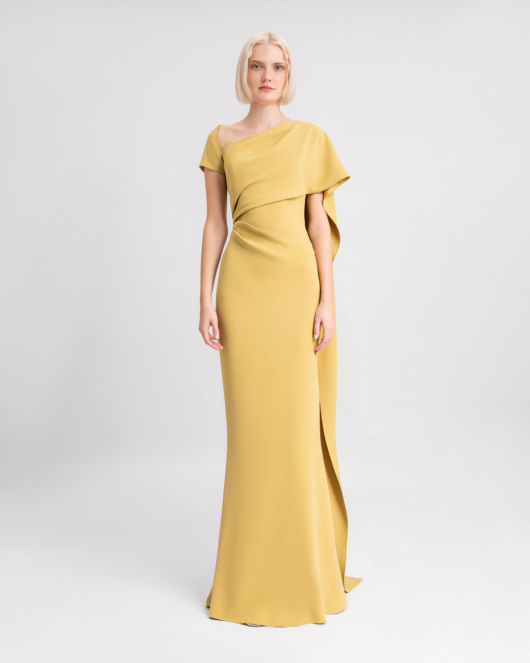 An asymmetrical neckline evening dress in mustard color featuring draping on the waistline and a long ruffled cape-like sleeve.