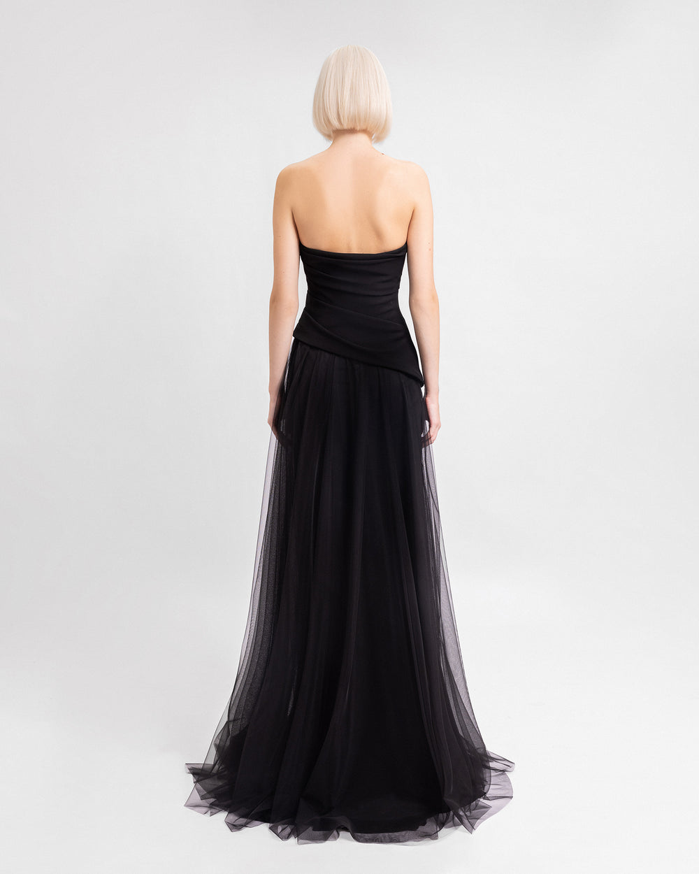 The back of a strapless black evening dress with an asymmetrically draped corset and a tulle skirt with a slit on the side.