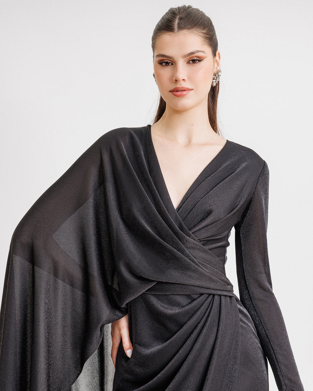 A close-up of a An asymmetrical V-neckline black evening dress with draping, a long sleeve from one side, and a cape-like sleeve on the other side.