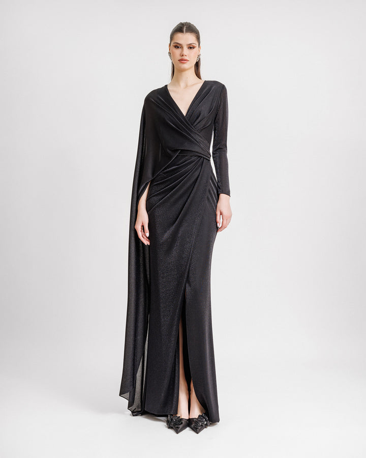 An asymmetrical V-neckline black evening dress with draping, a slit on the side, a long sleeve from one side, and a cape-like sleeve on the other side.