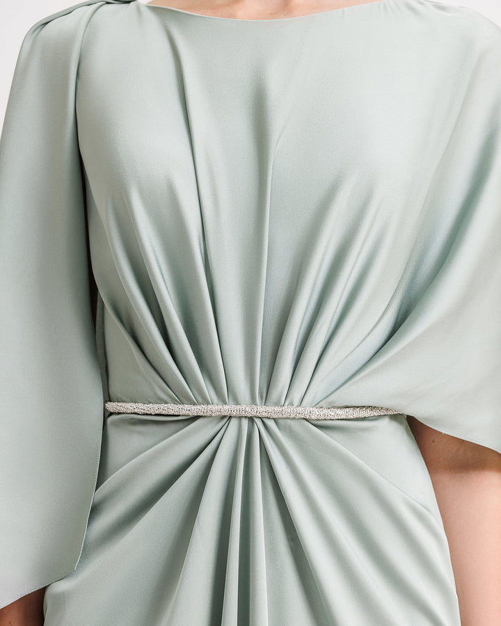 A close-up of a draped evening dress in teal satin with a beaded waist and asymmetrical cape-like sleeves.