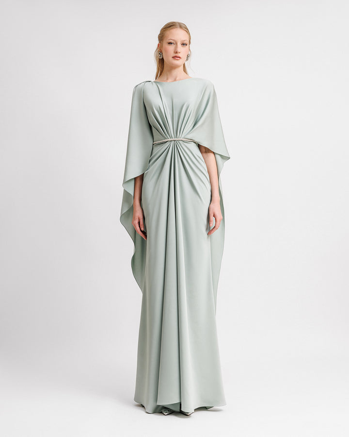 A draped evening dress in teal satin with a beaded waist and asymmetrical cape-like sleeves and back.