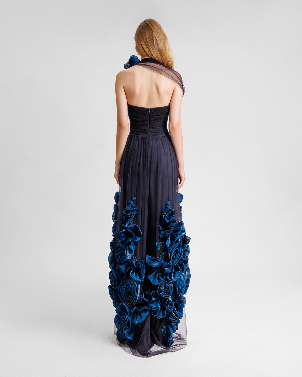 The back of a flared evening dress in navy draped tulle with an open back and a floral draping on the shoulder and hemline.