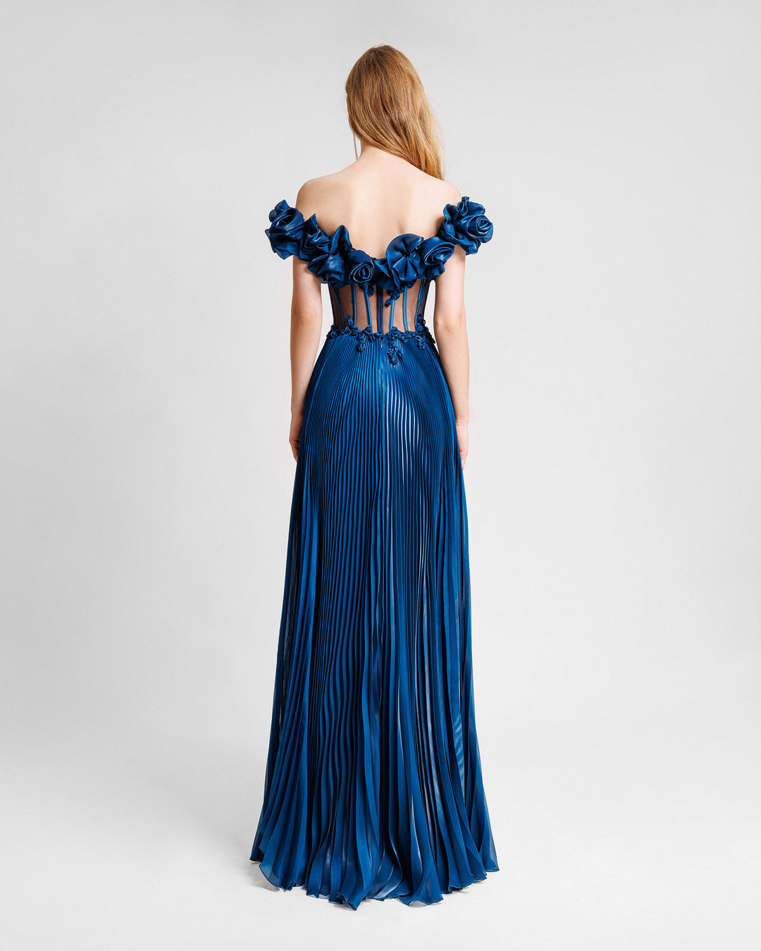 The back of a petrol evening dress with off-the-shoulders floral draping, featuring a see though corset and a pleated skirt.