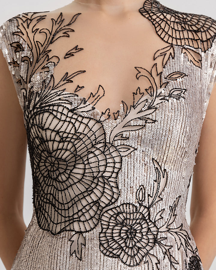 A close-up of an embroidered sequins slim-cut evening dress in greige color with a black beaded floral design, a V-neckline, and a slit on the side.