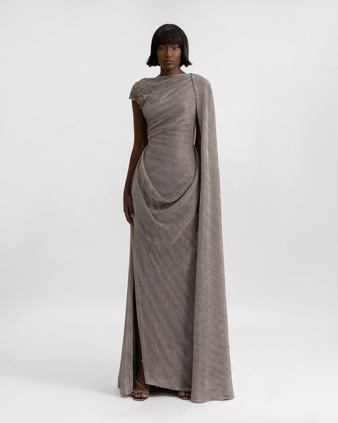 An asymmetrical draped dress in grey metallic jersey with a cape-like floor length sleeve, beadings on the shoulder, and a slit on the side.