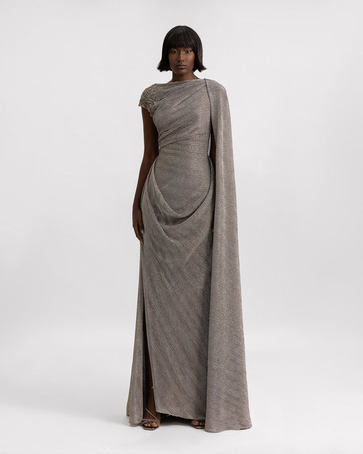 An asymmetrical draped dress in grey metallic jersey with a cape-like floor length sleeve, beadings on the shoulder, and a slit on the side.