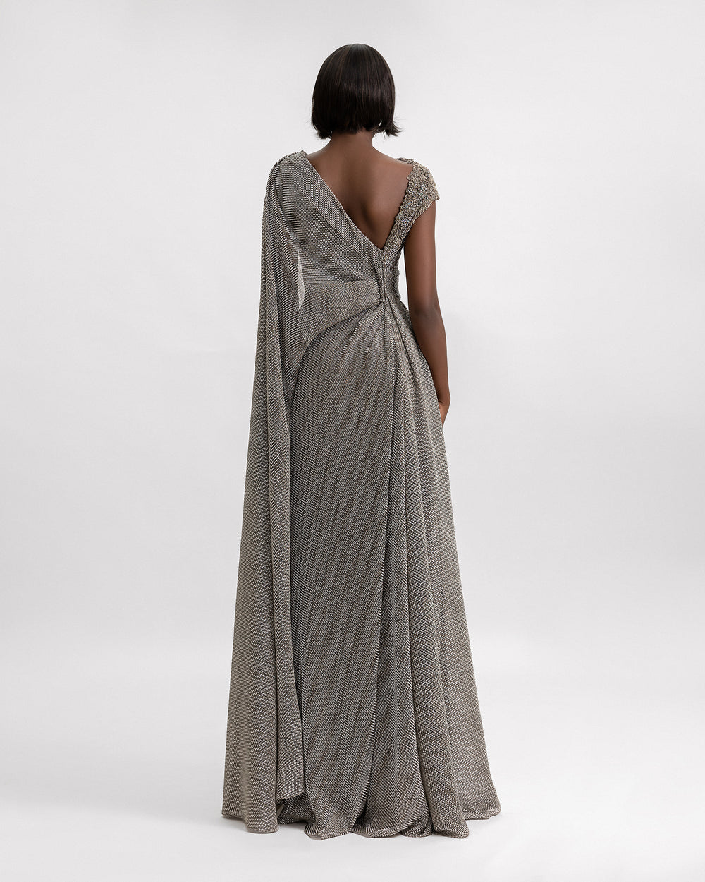 An asymmetrical v-shaped open back draped dress in grey metallic jersey with a cape-like floor length sleeve, and beadings on the shoulder.