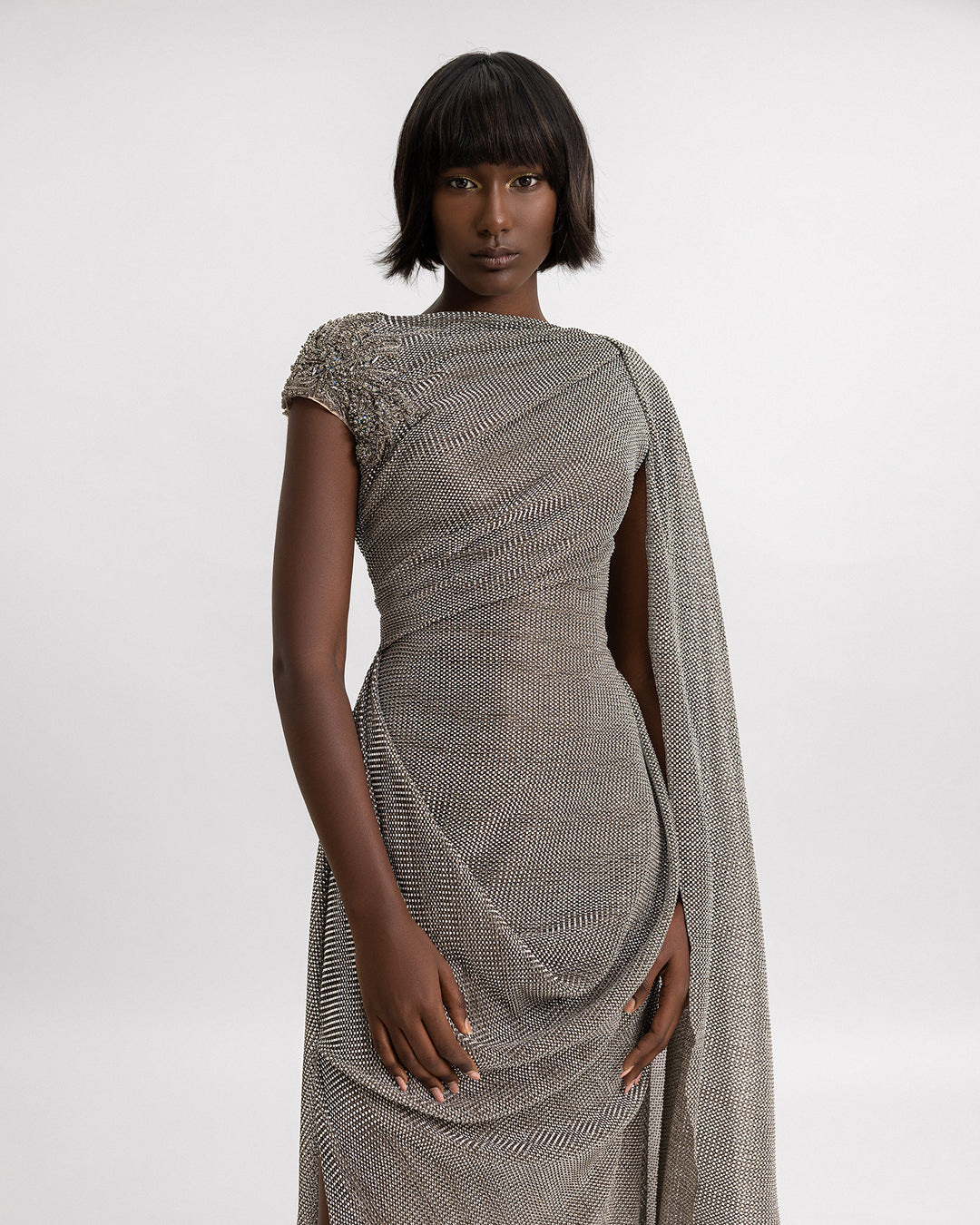 A close-up of an asymmetrical draped dress in grey metallic jersey with a cape-like floor length sleeve, beadings on the shoulder, and a slit on the side.