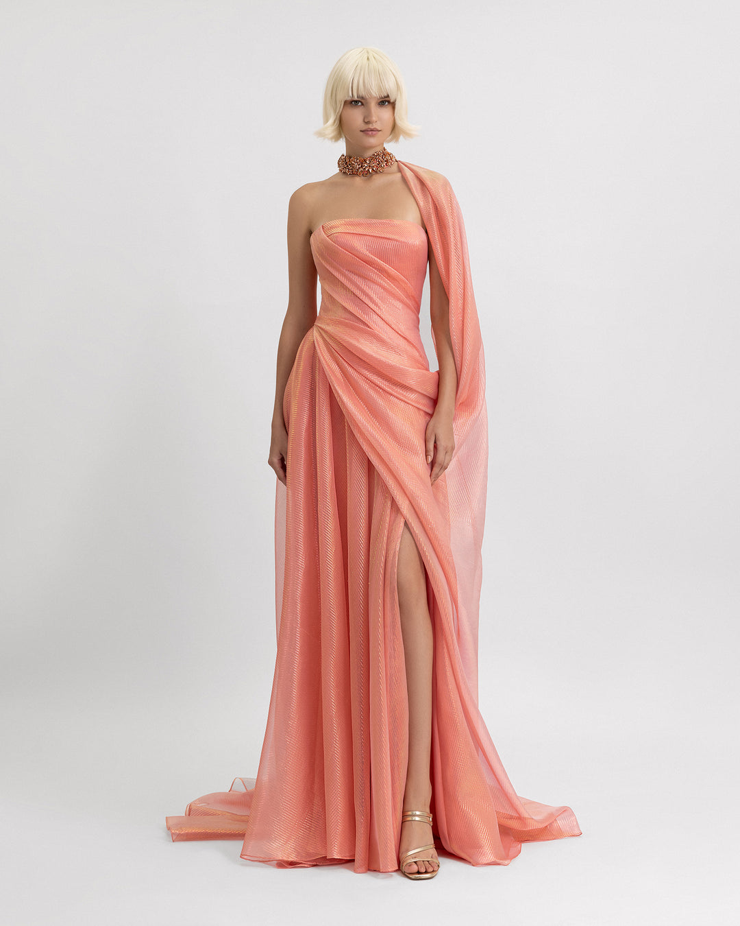 A strapless draped evening dress in coral color featuring a beaded collar, a draped cape-like detail, and a slit on the side.