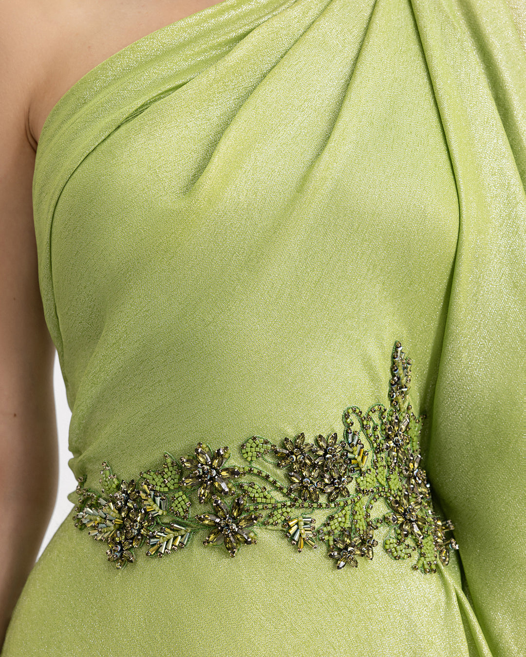 A close-up of a one-shoulder green evening dress with draping details and beadings on the waistline.