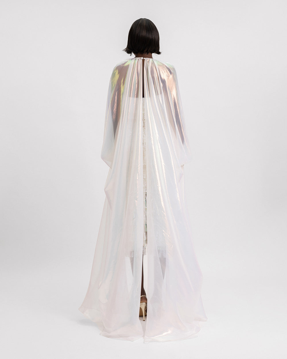 The back of a sequins tasseled ivory short dress featuring a chiffon irisé draped long cape with a slit at the back.