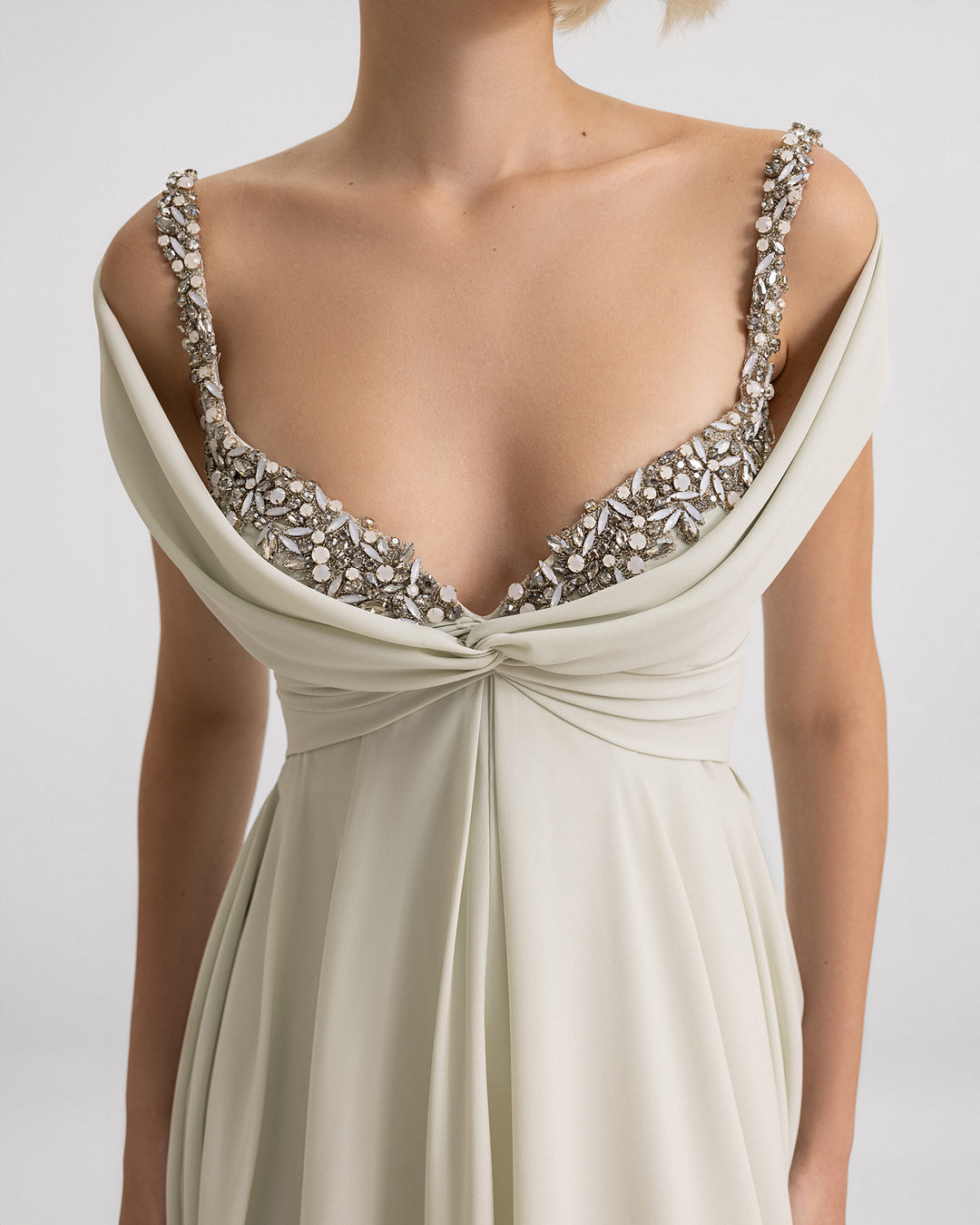 A close-up of a long evening dress in pale sage featuring off-the-shoulders draping and a beaded neckline.