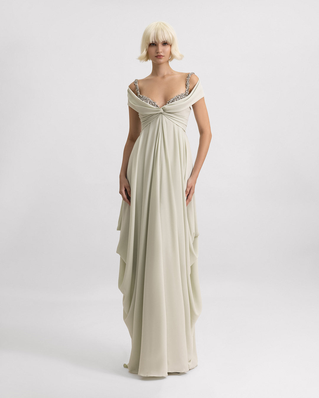 A long evening dress in pale sage featuring off-the-shoulders draping, beaded neckline, and draped skirt on the sides.