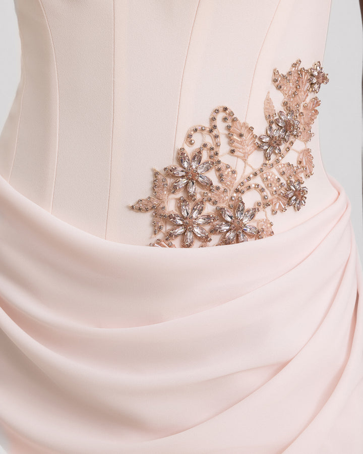 A close-up of a light pink evening dress featuring a structured corset, beading details on the waist, and a draped skirt.